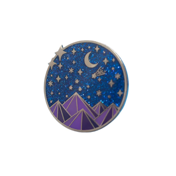 A colorful enamel pin with a fantasy court theme, featuring a starry night sky and mountains, accompanied by a butterfly clasp and placed on a blue background.