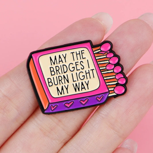 'May the bridges I burn light my way' matchbox enamel pin held in hand